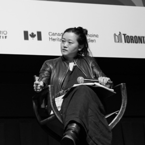 Woman on stage sitting cross-legged in a chair with a microphone on lap